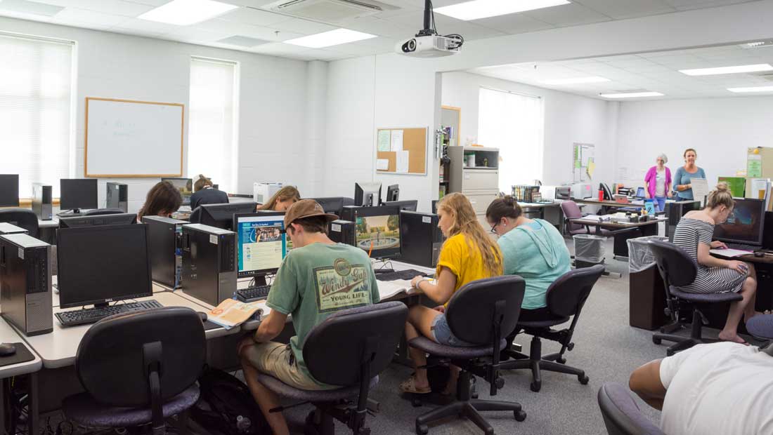 Students working at desktop computers in a college math, reading, and writing center with instructors in the background.